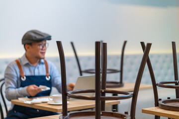 Wall Mural - Asian man coffee shop owner sitting at table and calculating shop finance bill on laptop computer after closed. Stressed male small business restaurant entrepreneur counting payment and income bills.