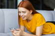 © kucherav - Smiling happy young woman with red hair texting on mobile phone lying on the couch at home