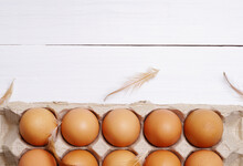 Top View Of Raw Organic Chicken Eggs In Egg Carton With Chicken Feather On White Wooden Background.