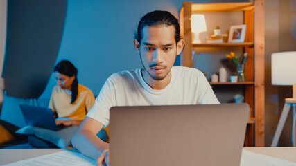 Poster - Freelance asia couple man and woman in casual feel serious focus on work separate laptop computer at night, hasband sit in front on table wife sit behind on couch at house. Work from home concept.