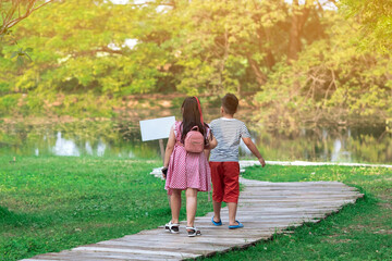 Wall Mural - Back view of Asian young girl and boy walking together on pathway through green garden. Sister and brother walking together in park. Happy family spending time together outside in green nature.