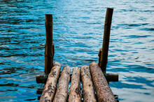 Wooden Bridge On The Background Of Blue Water. A Bridge Made Of Logs On The Lake.