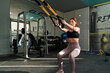© daniel - young latina woman performing squats with TRX inside the gym