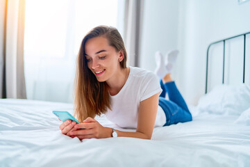 Wall Mural - Young happy smiling joyful millennial girl lying on a bed and using phone for reading news online, browsing web, social networking, chatting and watching movie at home