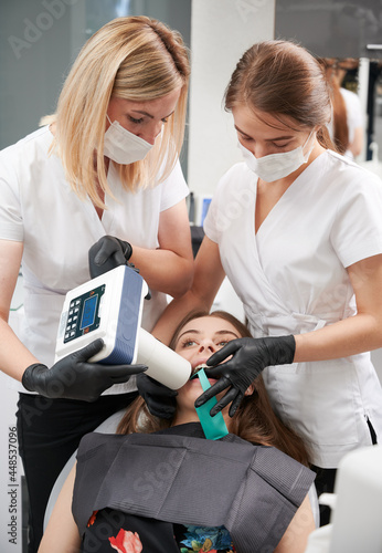 Doctors using portable dental x-ray machine while examining patient teeth in dental office, performing intraoral scanning with modern equipment. Concept of digital dentistry and intraoral radiography.