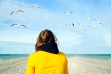 Back View Girl Wearing Yellow Jacket On The Shore With Seagulls At Sky. Girl Traveller Relaxing On The Beach And Feeds The Birds.