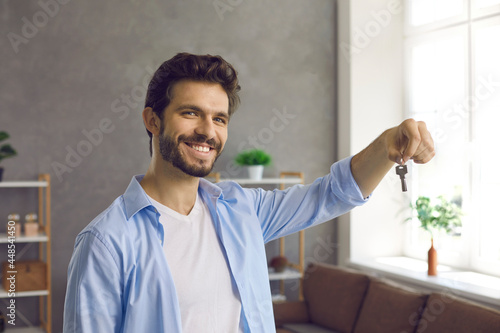 Happy handsome Caucasian young man looking at camera, smiling and showing us keys to his new home. Portrait of first time buyer, house owner, or apartment renter. Concept of buying your own property