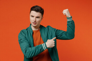 Wall Mural - Strong sporty fitness smiling young brunet man 20s wears red t-shirt green jacket pointing on biceps muscles on hand demonstrating strength power isolated on plain orange background studio portrait