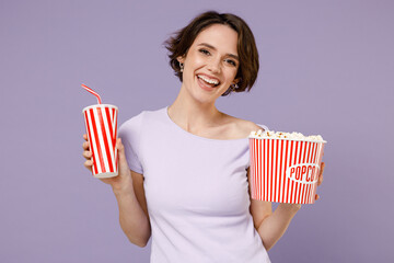 Poster - Young fun smiling happy woman 20s with bob haircut wearing white t-shirt hold takeaway popcorn bucket soda cola fizzy water in plastic cup isolated on pastel purple color background studio portrait