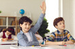 © Studio Romantic - Smart schoolboy sits at a desk next to his classmate in class with his hand raised, wanting to give the right answer or participate. Education, primary school, learning and people concept.