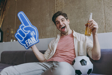 Wall Mural - Young overjoyed winner fun man 20s fan in casual clothes cheer up support football sport team with soccer ball drink beer fan foam glove finger up watch tv live stream sit on grey sofa indoor at home