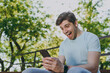 © ViDi Studio - Overjoyed exultant young happy man 20s in blue t-shirt sit on bench using mobile cell phone chat online scream rest relax in sunshine spring green city park outdoor on nature Urban leisure concept