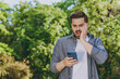 © ViDi Studio - Young confused shocked man 20s in blue casual shirt walk use mobile cell phone rest relax in spring sunshine forest green city park go down alley outdoors on nature. Urban lifestyle leisure concept