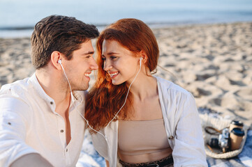 Close up happy young couple two family man woman in white clothes hug sitting sand listen to music headphones rest together at sunrise over sea beach ocean outside seaside in summer day sunset evening