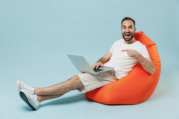 Wall Mural - Full length young smiling happy fun man in casual white t-shirt sit in bag chair hold use work on laptop pc computer rest relax isolated on plain pastel light blue color background studio portrait