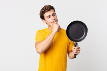 Wall Mural - young handsome man thinking, feeling doubtful and confused and holding a cook pan