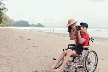 Wall Mural - Happy disabled teenage boy smile face on wheelchair holding a camera with mother, Activity outdoors with family on the beach background, People having fun and diverse people concept.