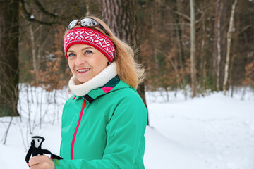 Portrait of elderly woman standing with nordic walking poles in winter forest, resting after exercise and looking at camera outdoors with smile.