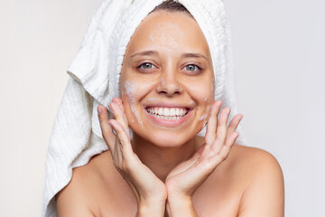 Wall Mural - A young beautiful caucasian smiling woman with a white towel on her head after a shower cleanses her face with foam for washing on a white background. Skin care, cosmetology. Morning routine. Close up