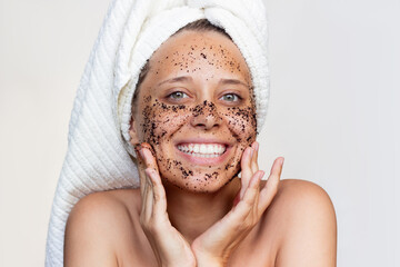 Wall Mural - A young beautiful caucasian smiling woman with a white towel on her head after a shower scrubs the face with a coffee mask on a white background. Skin care, cosmetology. Morning routine. Close up