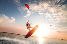 Sports Shot Of A Young Caucasian Woman In A Wetsuit Doing A Trick In The Air Against The Backdrop Of A Sunset In The Sea. Kitesurring Girl Athlete Flies On A Kite With A Board