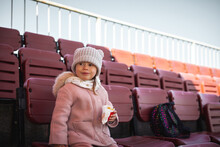 Cute Little Caucasian Girl Eating French Fries At The Stadium While Watching A Football Game. Beautiful Little Girl In A Stylish Outfit
