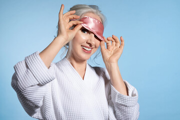 Joyful elderly female in sleeping mask, bathrobe, studio portrait on blue background with blank space. Positive rested mature lady take off eyemask. Healthy feel and refresh after night nap