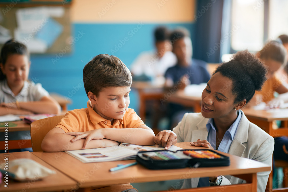 Happy African American teacher assisting her elementary student in classroom. - obrazy, fototapety, plakaty 