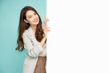 Cheerful Young Asian Businesswoman Is Standing Behind The White Blank Banner Or Empty Copy Space Advertisement Board On Green Background, Looking At Camera
