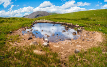 Green Swampy Plateau And A Blue Puddle Against The Background Of Distant Hilly Mountains. A Panorama Of A Pure Green Landscape.