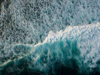 Drone photo of The dramatic lapping of the waves in the ocean produces white foam. The sea water of the pond is blue and contrasts with the color of the foam