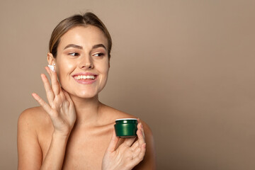 Wall Mural - Young woman applies moisturizer to her face and smiles. Skin care concept