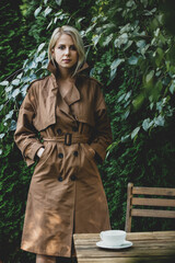 Sticker - Beautiful woman in coat stay next to table with cup of coffee in a garden with trees on background