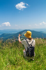 Wall Mural - Hipster traveler in a hat and backpack, resting in nature and using the Internet on a smartphone. A tourist girl photographs a panorama of the mountains by a mobile phone. Mountain beautiful landscape