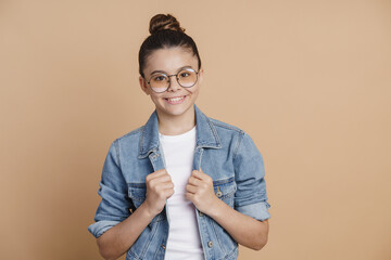 Wall Mural - Modern teenage girl dressed in casual style, denim jacket and jeans on a brown background. Cute little girl smiles, wears glasses and a bunch of hair on her head