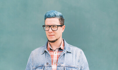 Close-up street portrait guy with blue hair in jeans jacket on blue wall background.