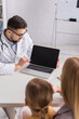© LIGHTFIELD STUDIOS - Pediatrician looking at laptop with blank screen near blurred child and parent in hospital