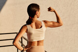 Back view portrait of stunned girl with dark hair and ponytail wearing sporty top standing with raised hand, showing her biceps and triceps, results of everyday workout.