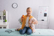 © LIGHTFIELD STUDIOS - Girl with pills and soft toy smiling at camera near stethoscope and electronic thermometer