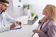 © LIGHTFIELD STUDIOS - smiling doctor writing prescription near woman touching sore throat during consultation