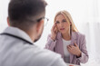© LIGHTFIELD STUDIOS - woman touching head and chest while talking to blurred doctor during consultation
