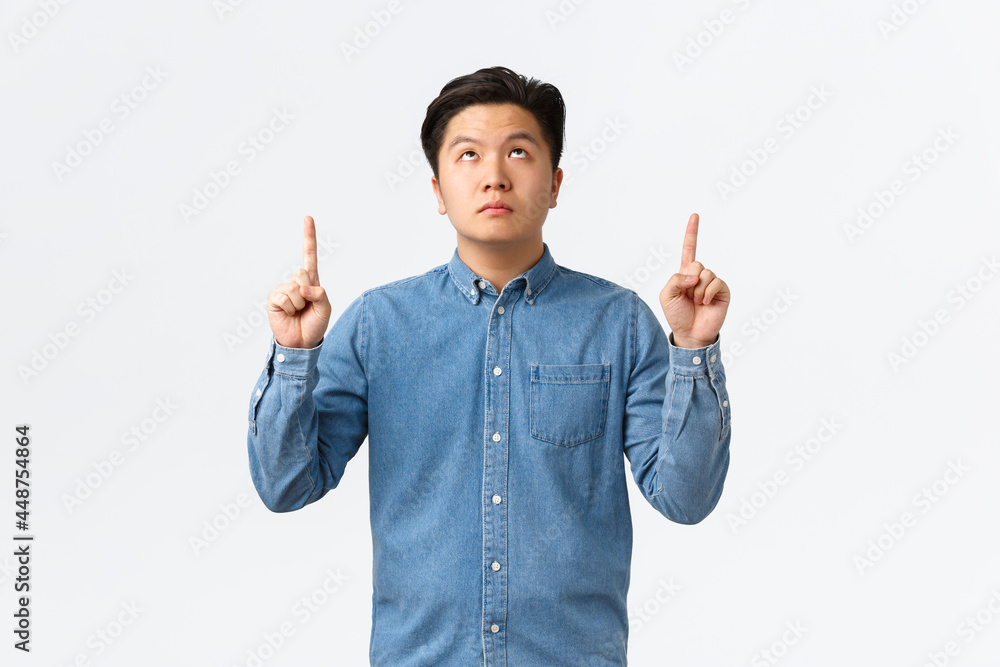 Unamused bored asian guy in blue shirt, looking and pointing fingers up ...
