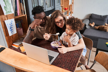 Wall Mural - Woman feeding with breakfast her lovely multiracial child while sitting in front of laptop