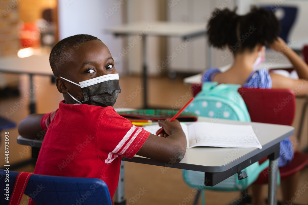 Portrait of african american boy wearing face mask sitting on his desk in class at elementary school - obrazy, fototapety, plakaty 