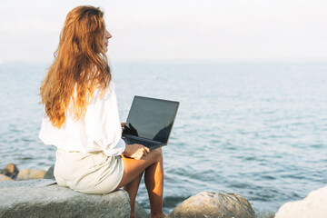 Young carefree beautiful freelance businesswoman with long hair in a white shirt working at laptop on seashore on sunrise