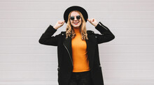 Fashionable Portrait Of Stylish Happy Smiling Young Woman Having Fun Female Model Posing Wearing A Black Coat, Round Hat On City Street On Gray Background
