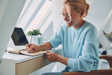 Wall Mural - Beautiful young smiling woman writing something in note pad while working at home