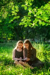 Wall Mural - Two schoolgirls in school uniforms are reading a book while sitting on the green grass in the park. Do homework