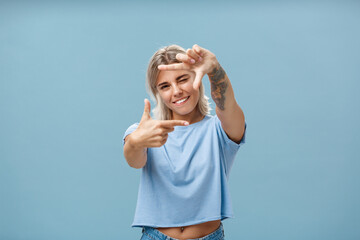 Be creative and happy. Portrait of charming enthusiastic female designer with blond hair and tattoo on arm winking and smiling joyfully making frame gesture as if measuring space over blue background