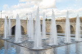 Fototapeta Most - Decorative jets of water rising vertically in the park of Madrid Rio one day with clouds and with the stone bridge of Segovia in the background